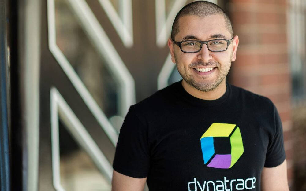 A smiling man wearing glasses and a Dynatrace t-shirt, standing in front of a modern building with geometric patterns